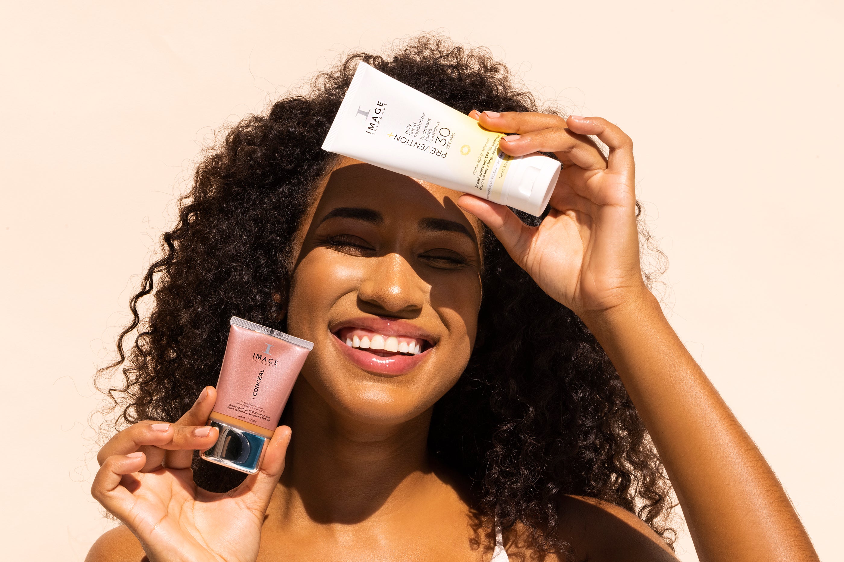 A woman smiling while holding two IMAGE Skincare products: Prevention + daily tinted moisturizer and a concealer